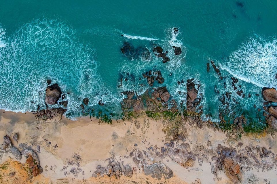 An aerial, top-down view of turquoise ocean waves washing over a rocky, sandy coastline.