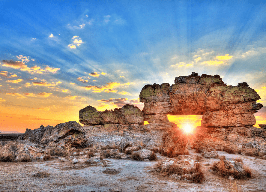 The setting sun shines through a natural arch in a large rock formation, with rays of sunlight radiating across a blue-yellow sky.