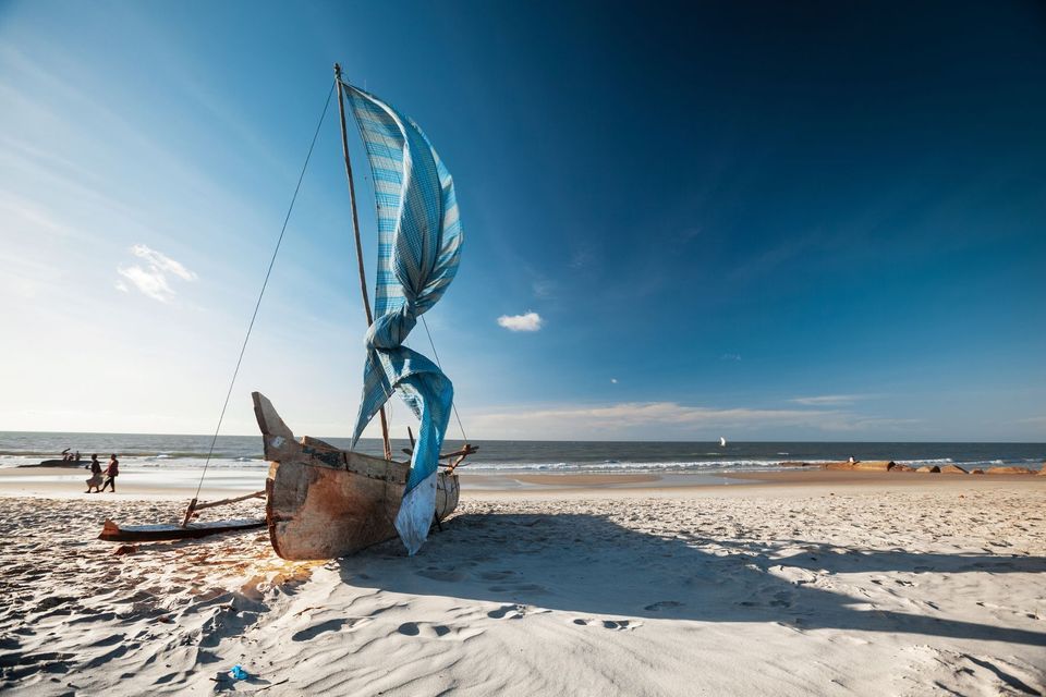 A wooden boat with a furled blue and white sail rests on a sandy beach next to the ocean under a clear blue sky.