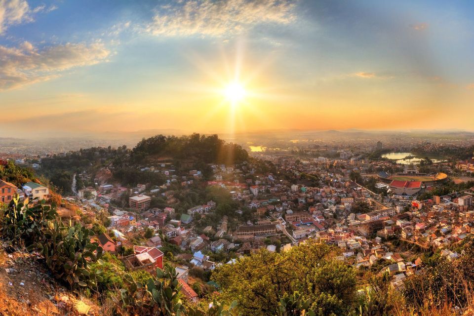 A panoramic view of a sprawling city built across rolling hills, with the sun setting brightly over the horizon.