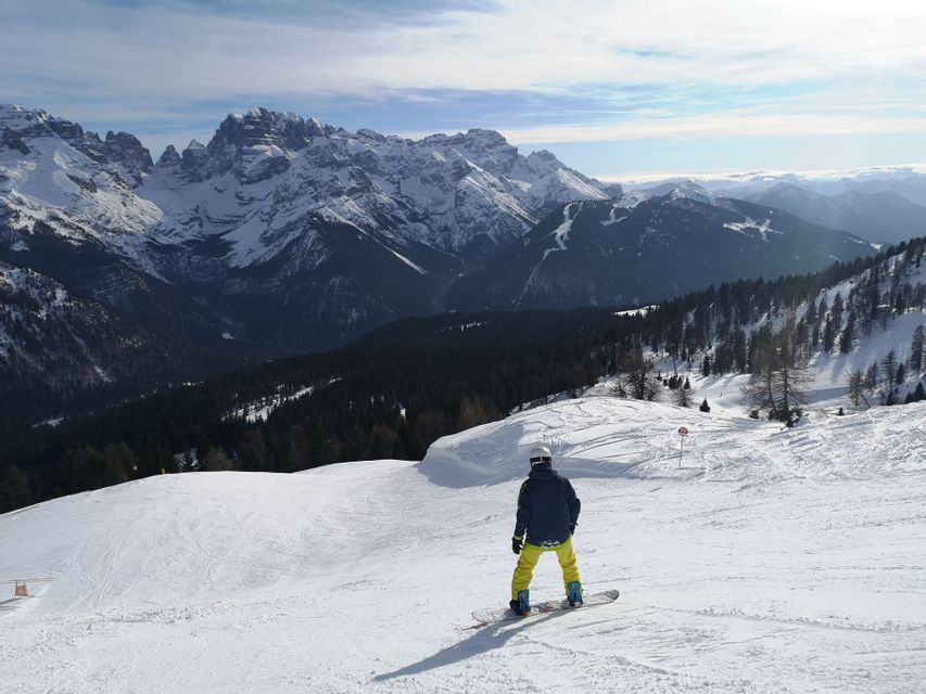 Uno snowboarder si ferma su un pendio innevato, ammirando una vasta catena montuosa coperta di neve sotto un cielo parzialmente nuvoloso.