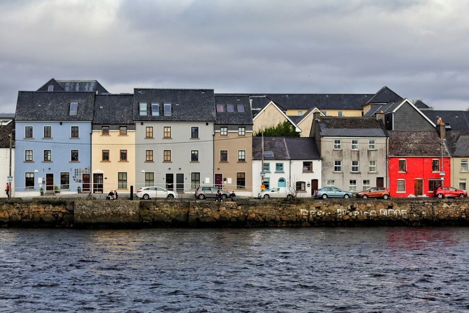 Une rangée de maisons colorées avec des voitures garées se dresse sur un quai en pierre au bord de l'eau sous un ciel nuageux.