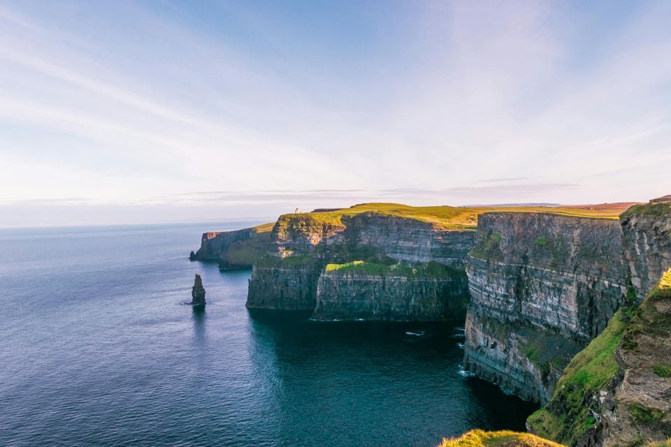 De hautes falaises rocheuses couvertes d'herbe verte surplombent une mer d'un bleu foncé avec un piton rocheux proéminent.
