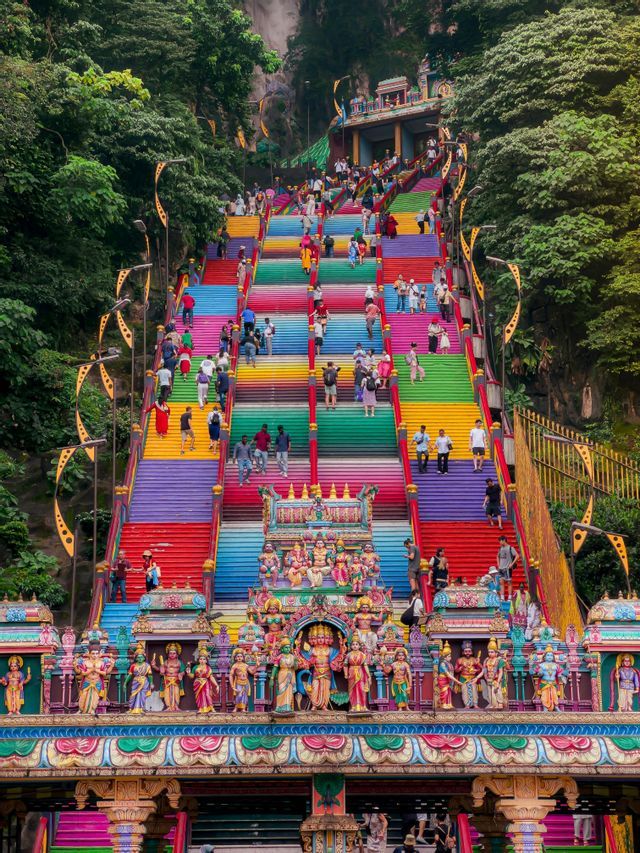 Eine Menschenmenge steigt eine steile, regenbogenfarbene Treppe zu einem Tempel an einem üppigen, grünen Berghang empor.