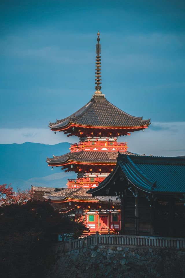 Una pagoda giapponese alta e rossa con più piani e tetti di tegole scure, sullo sfondo di un cielo blu e montagne distanti.