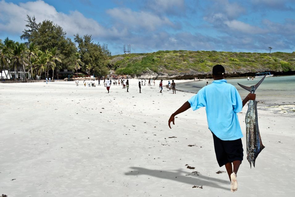 Un homme en chemise bleue traverse une plage de sable blanc, portant un grand poisson par la queue.