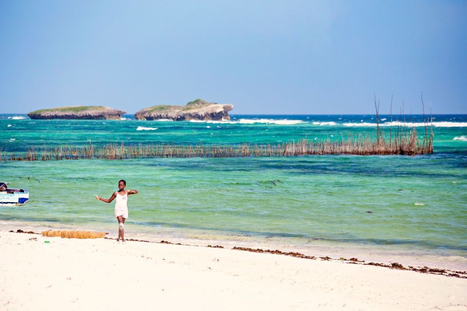 Une jeune fille en robe claire se tient sur une plage de sable blanc, au bord d'une eau turquoise, avec des îlots rocheux en arrière-plan.