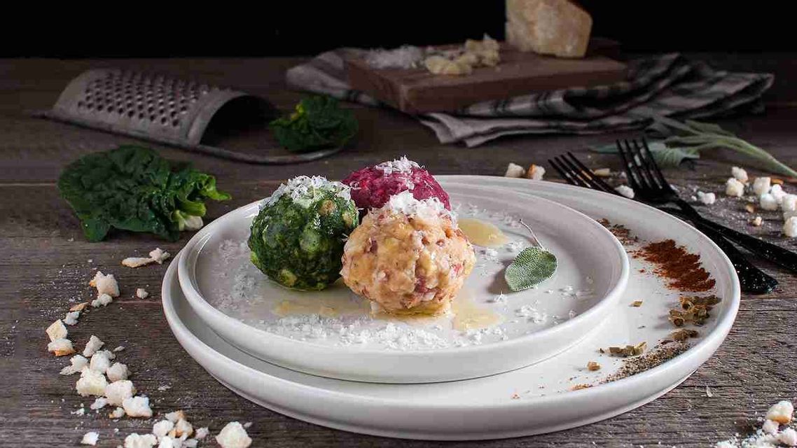 Un piatto di tre gnocchi colorati—verdi, rossi e beige—guarniti con formaggio grattugiato e una foglia di salvia su un tavolo di legno.