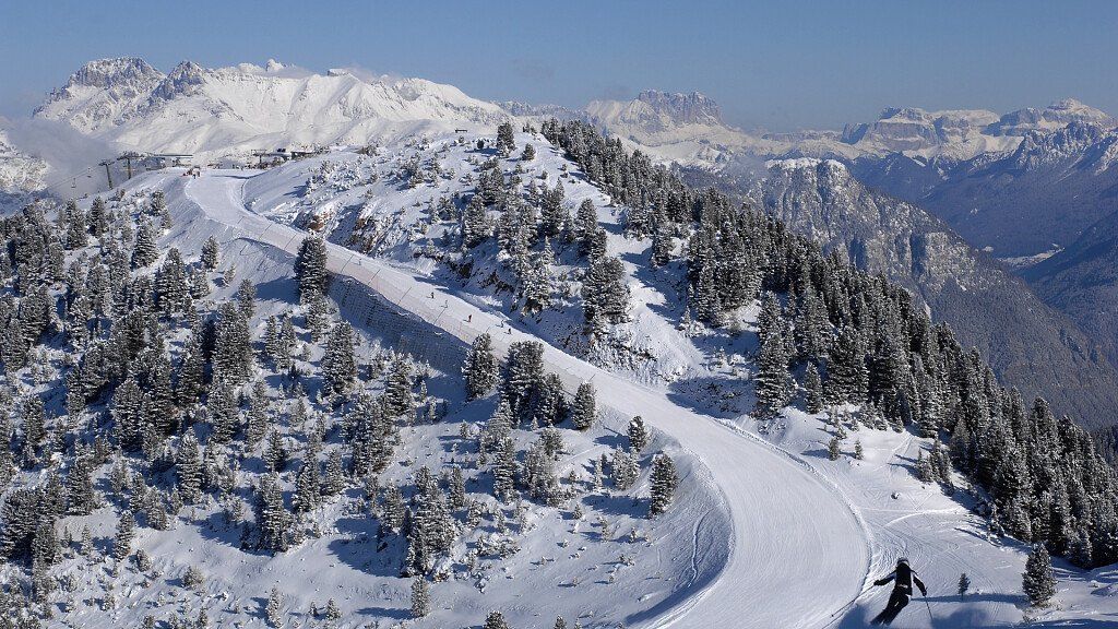 Uno sciatore scende una pista ampia e battuta, circondato da pini coperti di neve e vette montuose distanti.