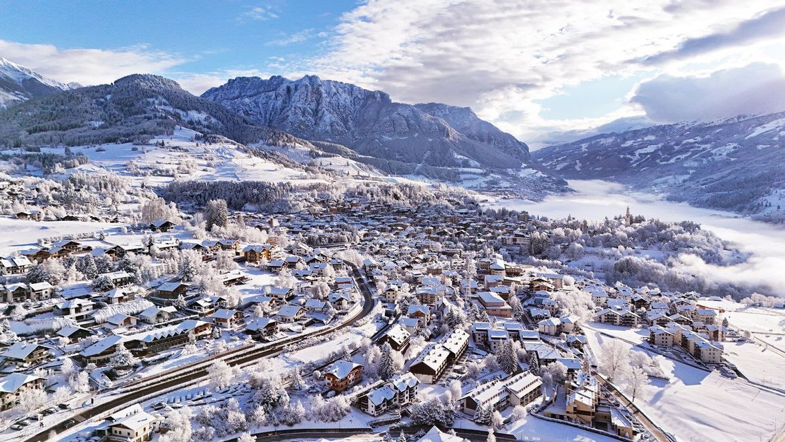 Una vista aerea di un villaggio con tetti innevati annidato in una valle montana sotto un cielo parzialmente nuvoloso.