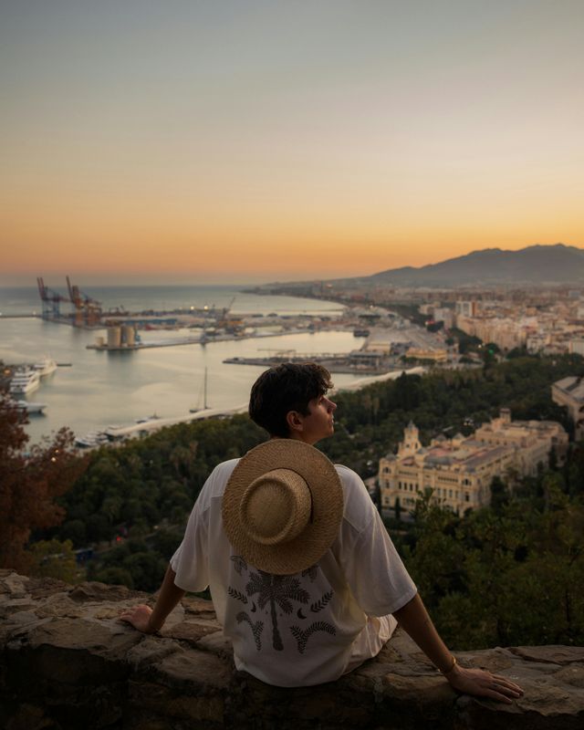 Una persona con un cappello di paglia sulla schiena siede su un muro di pietra, osservando una città costiera e il porto al tramonto.