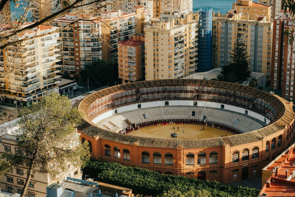 Una vista aerea di un'arena circolare per corride incastonata tra edifici cittadini e alberi, con il mare sullo sfondo.