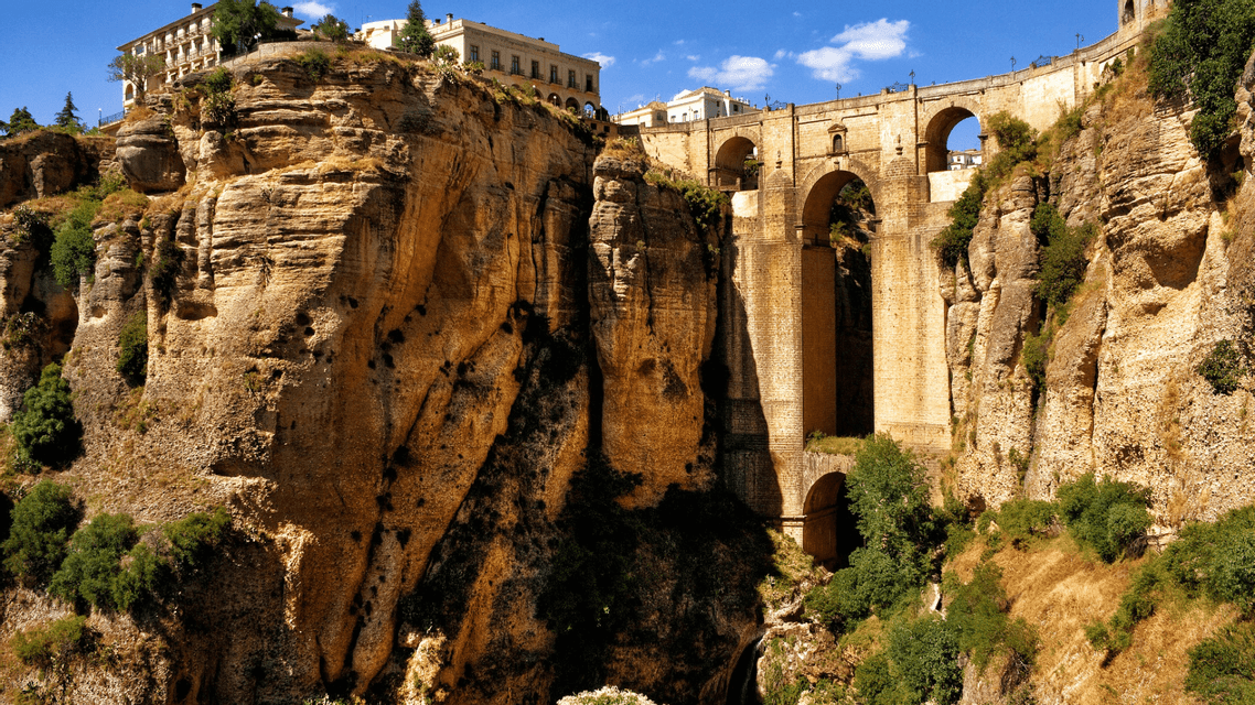 A large stone bridge with multiple arches spans a deep, rocky gorge with buildings perched on the clifftop under a blue sky.