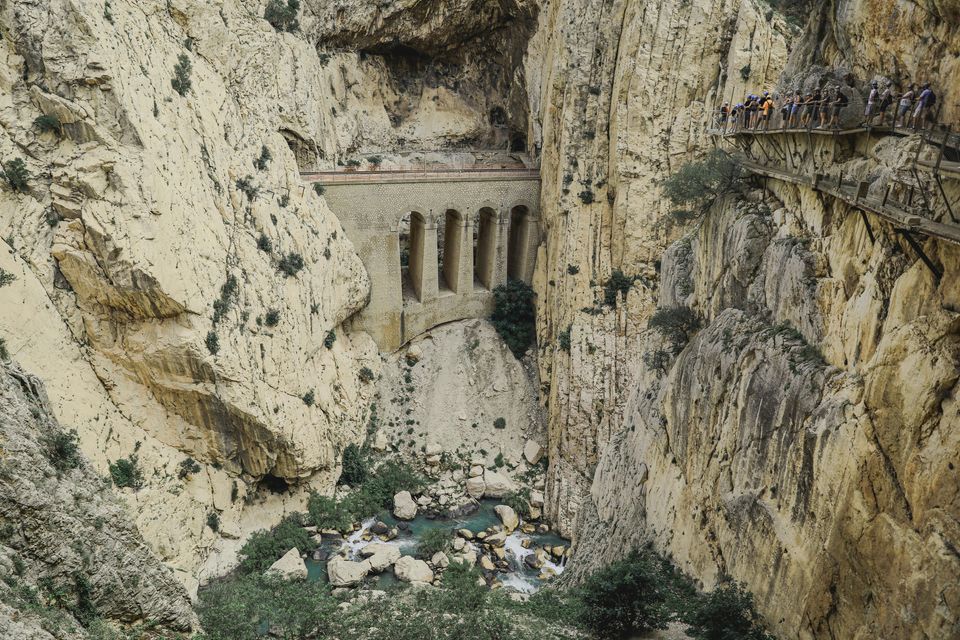 Un viaggio di gruppo WeRoad percorre uno stretto sentiero ricavato nella parete a strapiombo di un profondo canyon, con un acquedotto sottostante.