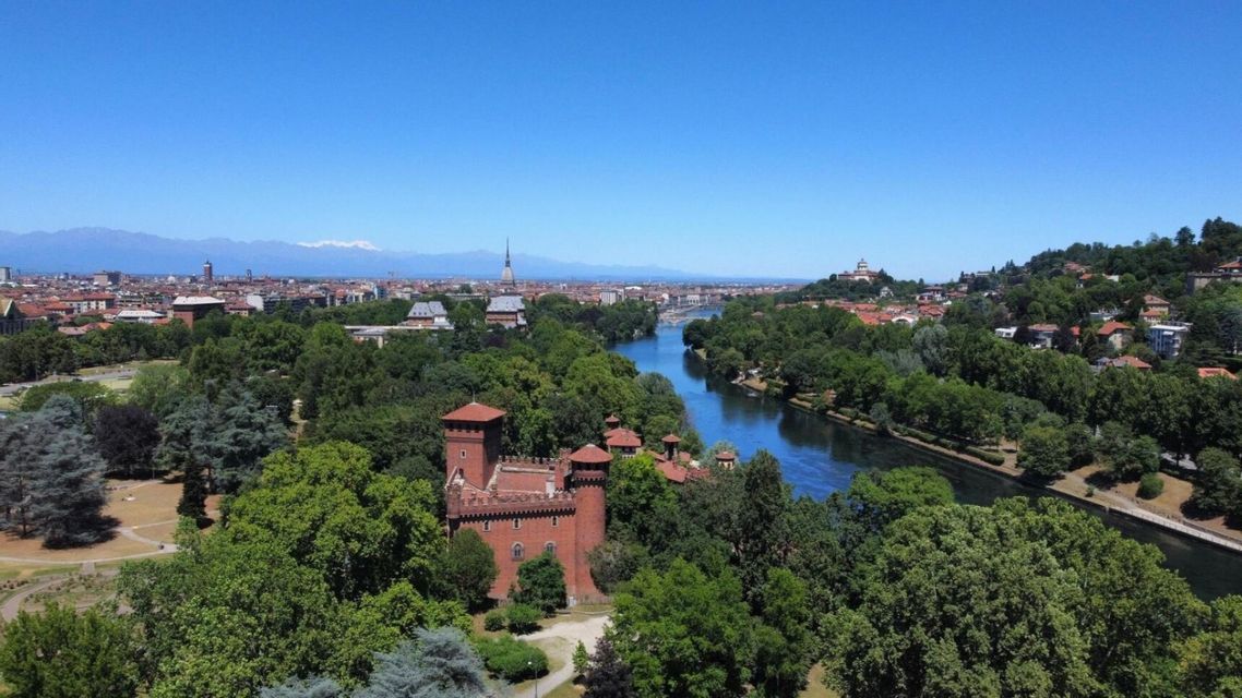 Vista aerea di un castello in mattoni rossi in un rigoglioso parco verde accanto a un ampio fiume, con uno skyline cittadino e montagne distanti.