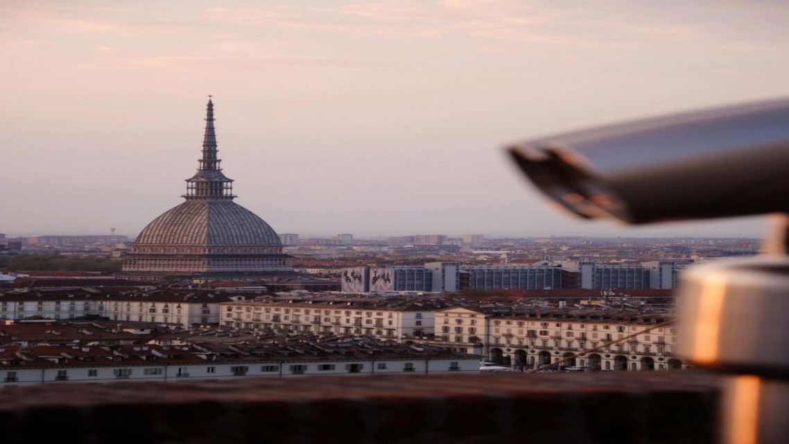Un paesaggio urbano con un imponente edificio a cupola e un'alta guglia, visto da un punto panoramico elevato con un telescopio in primo piano.