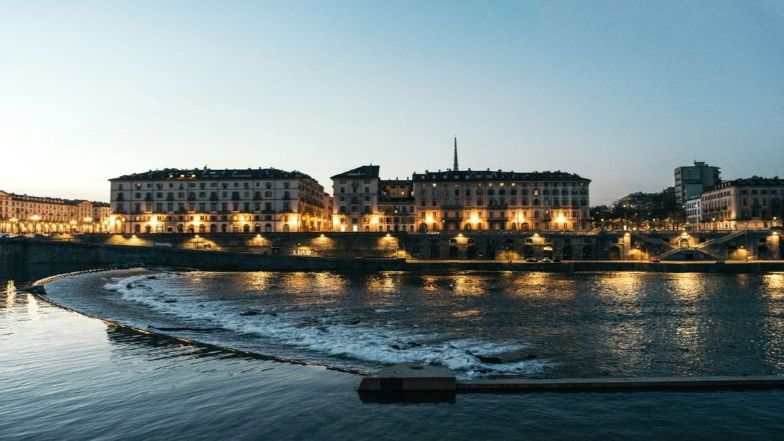 Una fila di edifici illuminati costeggia un fiume al tramonto, con le luci che si riflettono sull'acqua che scorre.