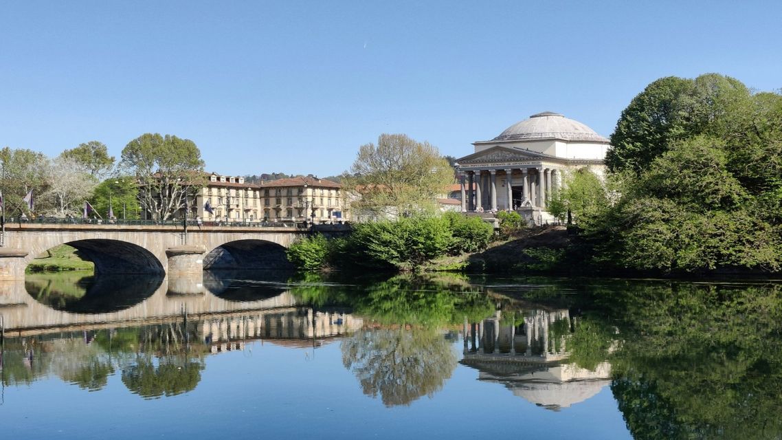 Un classico edificio a cupola e un ponte ad arco in pietra si riflettono nell'acqua calma di un fiume sotto un cielo azzurro limpido.