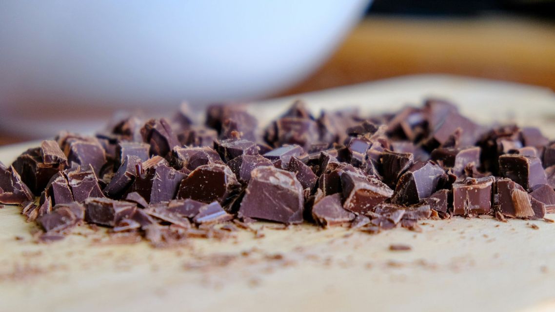 Un mucchio di pezzi e scaglie di cioccolato fondente tritato grossolanamente su un tagliere di legno chiaro.