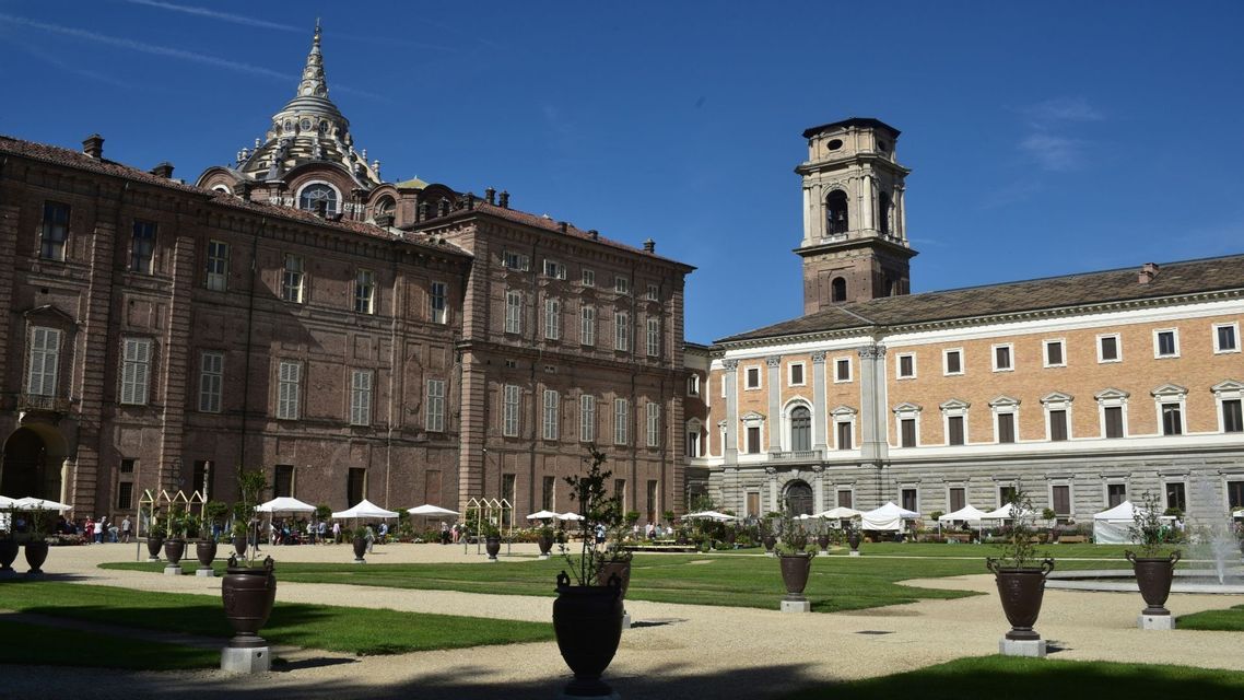 Edifici storici con una cupola prominente e un campanile si affacciano su un cortile soleggiato con prati, sentieri di ghiaia e grandi fioriere.