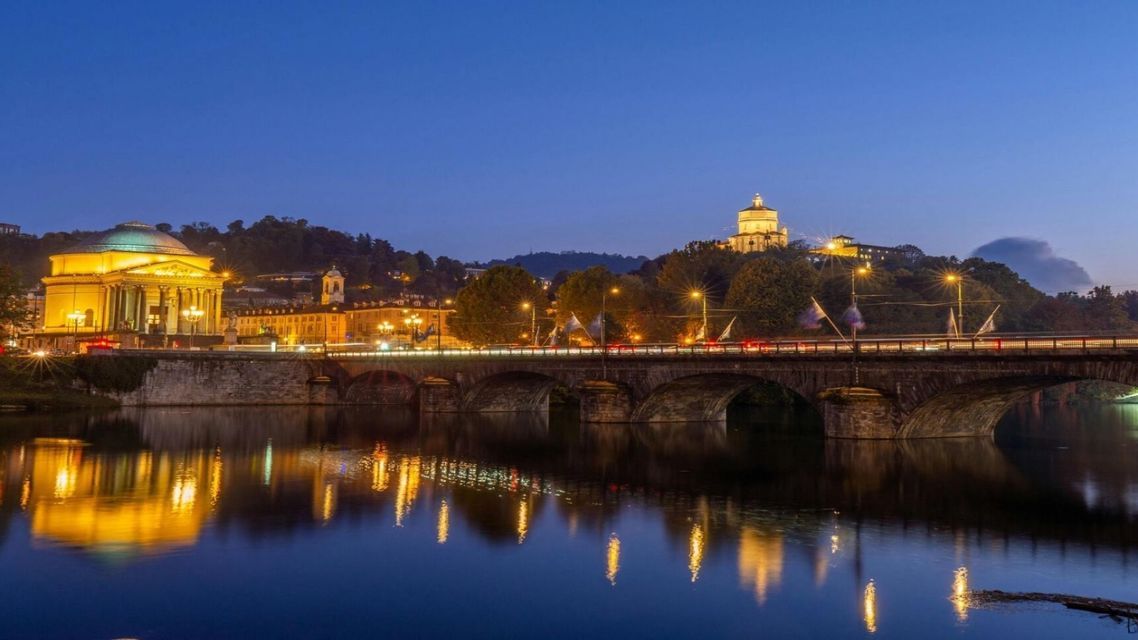 Un ponte ad arco in pietra e edifici storici illuminati al crepuscolo, con le loro luci che si riflettono sul fiume calmo sottostante.