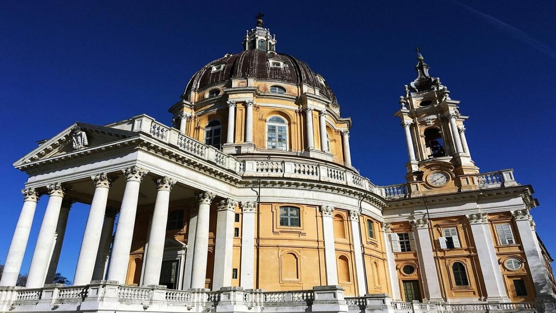 Veduta dal basso di un'ornata basilica barocca con un'ampia cupola centrale, colonne bianche e un campanile contro un cielo azzurro limpido.