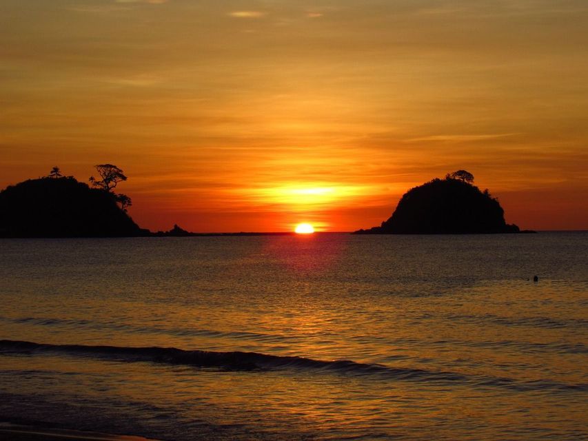 Il sole tramonta all'orizzonte tra due isole in silhouette, proiettando un bagliore arancione sul mare calmo.