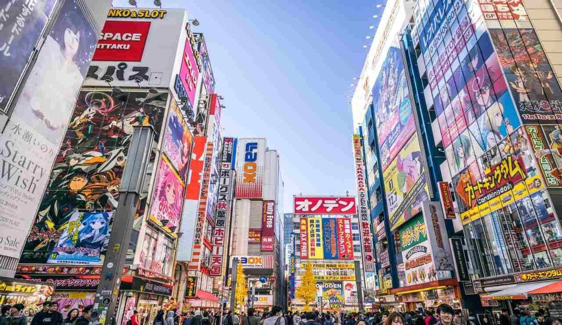 Una strada cittadina trafficata con edifici alti coperti da colorate pubblicità giapponesi e cartelloni in stile anime sotto un cielo limpido.