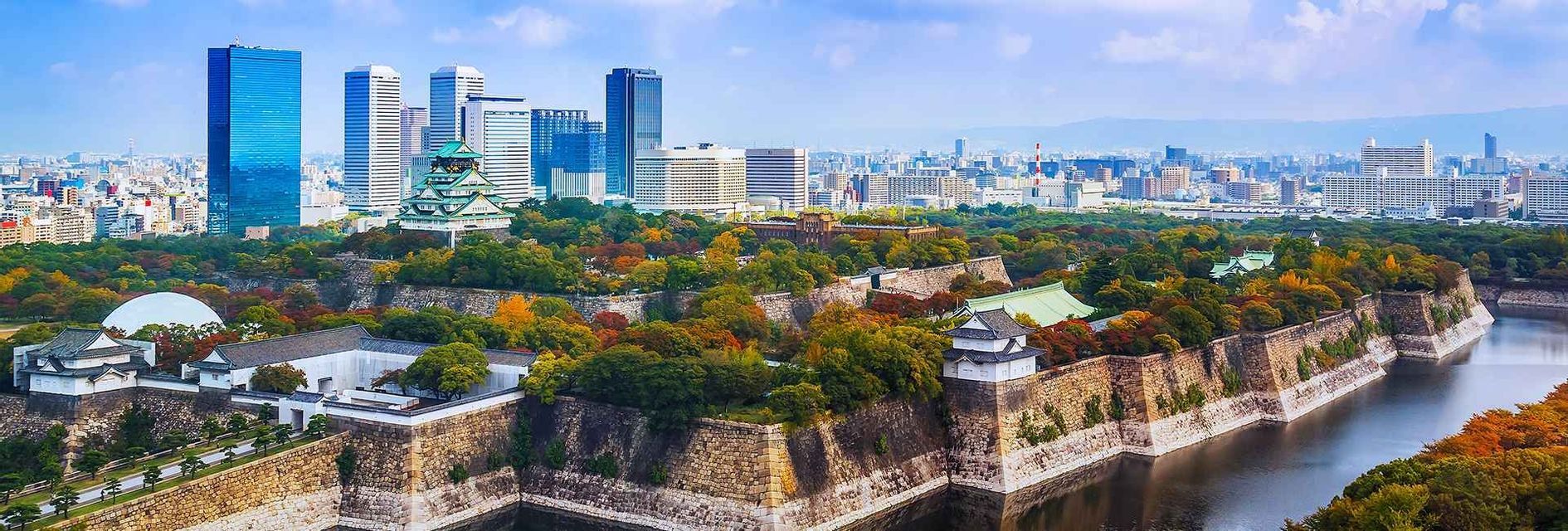 Veduta panoramica di un castello giapponese tradizionale con fossato, circondato da alberi autunnali e lo skyline di una città moderna sullo sfondo.