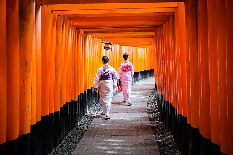 Due donne in kimono tradizionali percorrono un sentiero di pietra attraverso un tunnel di vivaci porte torii arancioni.
