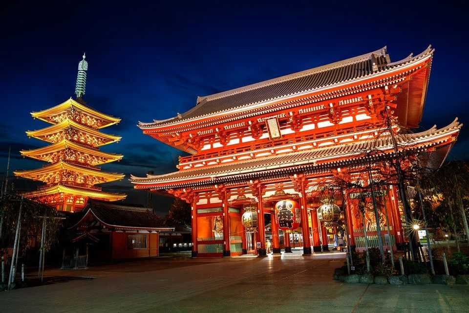 Un grande portale rosso del tempio e una pagoda a cinque piani sono illuminati da una luce dorata contro un cielo notturno blu intenso.