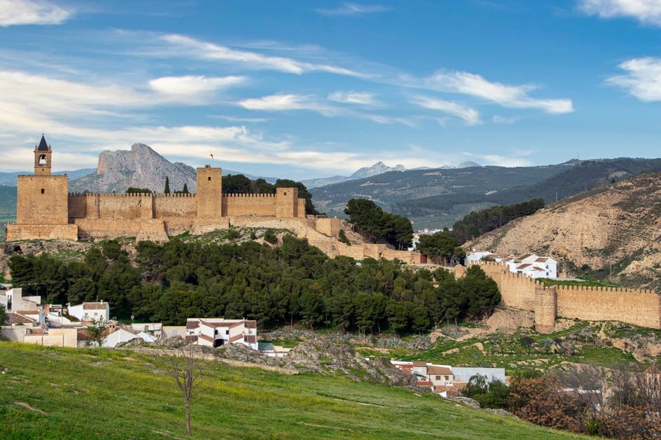 A large sand-colored fortress with a bell tower and battlements sits on a tree-covered hill above a village.