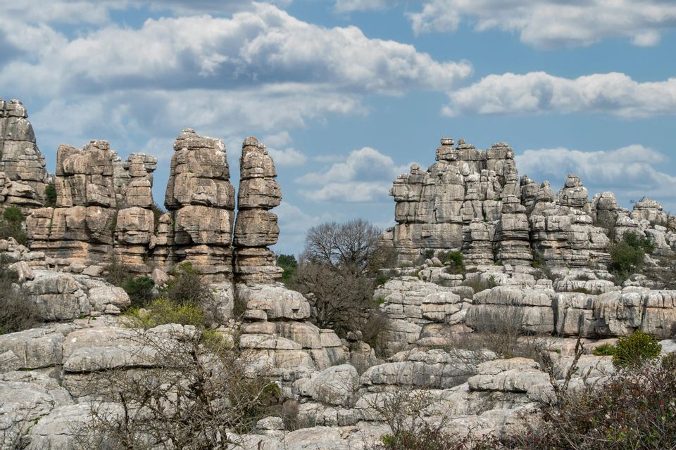 Tall, layered rock formations rise from a rugged landscape with sparse vegetation under a partly cloudy sky.