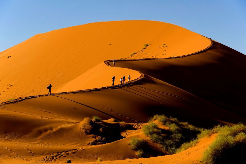 Un gruppo WeRoad cammina in fila lungo il sinuoso crinale di una vasta duna di sabbia arancione, contro un cielo azzurro limpido.