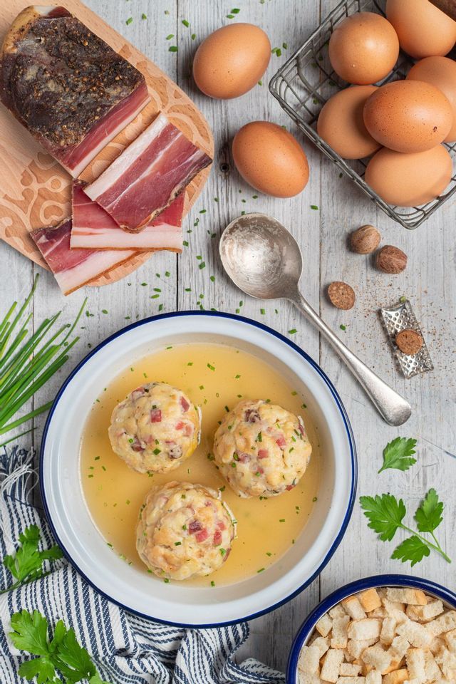 Una ciotola di canederli di speck in brodo su un tavolo di legno, circondata da ingredienti tra cui speck a fette, uova e cubetti di pane.