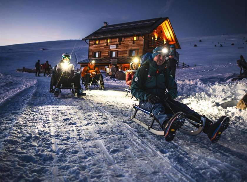 Un gruppo WeRoad si gode una slittata notturna con lampade frontali su un sentiero innevato fuori da una baita in legno.
