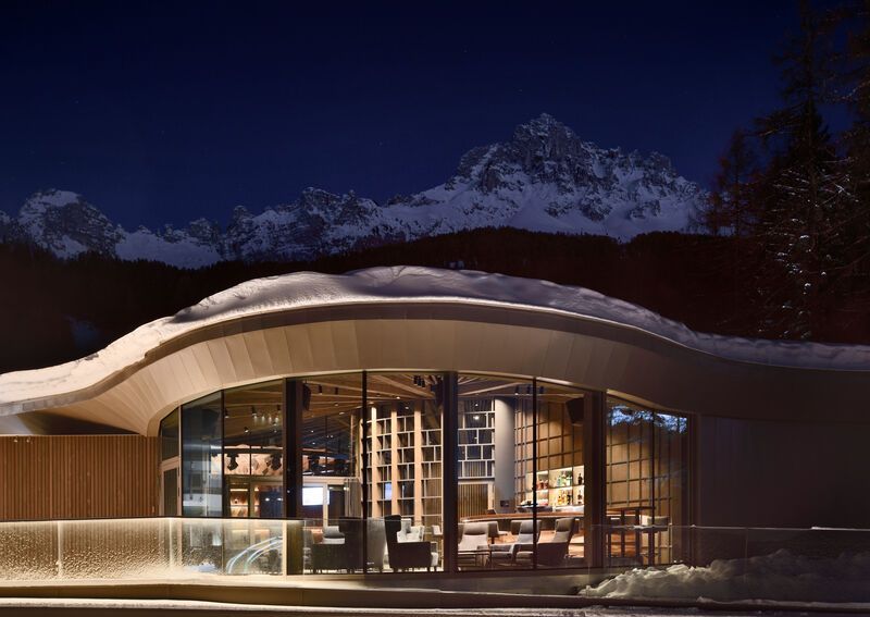 Un edificio moderno con tetto curvo e innevato è illuminato di notte, con imponenti montagne innevate sullo sfondo.