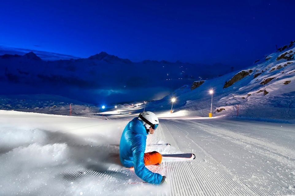 Uno sciatore con giacca blu e casco bianco siede su una pista da sci appena battuta di notte, illuminata dai fari della pista.