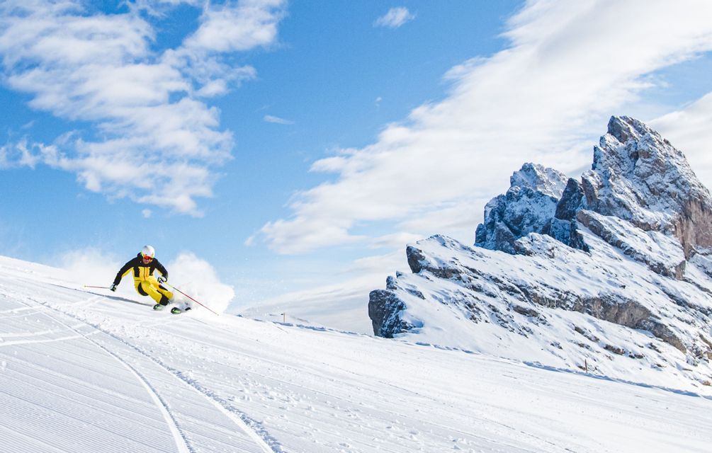Una persona con una tuta da sci gialla e nera scia su una pista battuta con montagne innevate sotto un cielo azzurro.