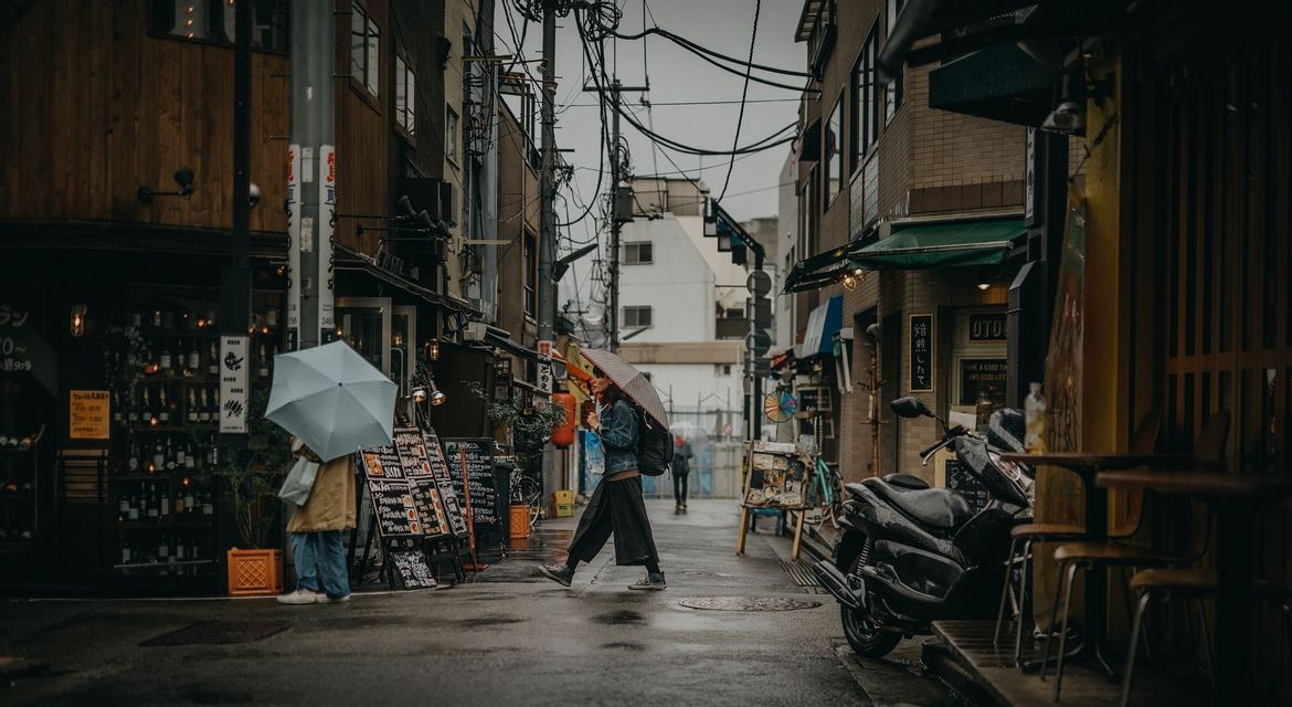 Persone con ombrelli camminano attraverso un vicolo stretto e bagnato di città, fiancheggiato da negozi e fili aerei aggrovigliati.