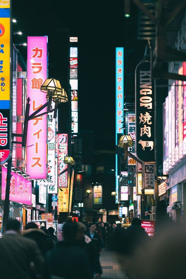 Una folla di persone cammina lungo una strada stretta di notte, illuminata da alte insegne al neon luminose con caratteri giapponesi.