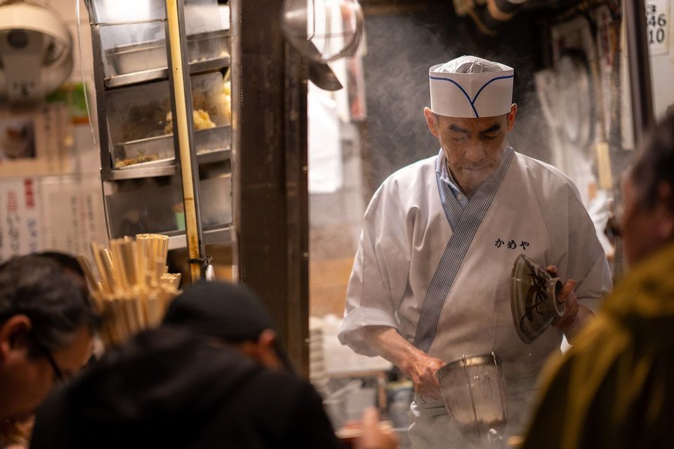 Un anziano chef giapponese in uniforme bianca e cappello di carta prepara il cibo con un colino di metallo in una cucina piena di vapore.