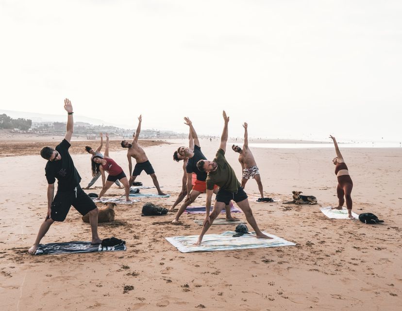 Un viaggio di gruppo WeRoad pratica yoga in posizione del triangolo su una spiaggia sabbiosa, con diversi cani che riposano sulla sabbia.