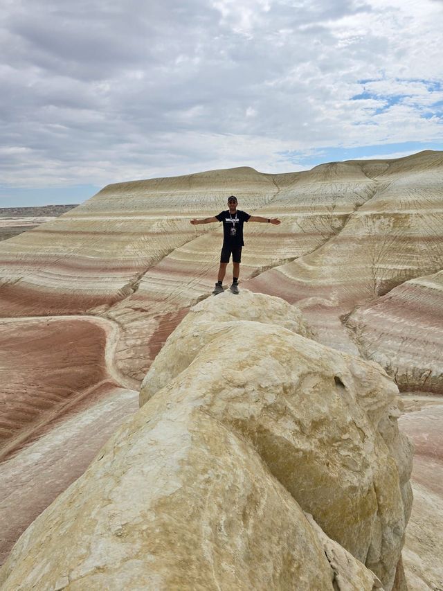 A man stands with arms outstretched on a rock formation, overlooking a canyon of colorful, striped hills under a cloudy sky.