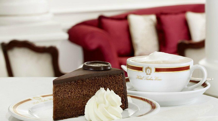 Una fetta di torta al cioccolato con medaglione e panna montata è servita accanto a un cappuccino schiumoso su un tavolo bianco.