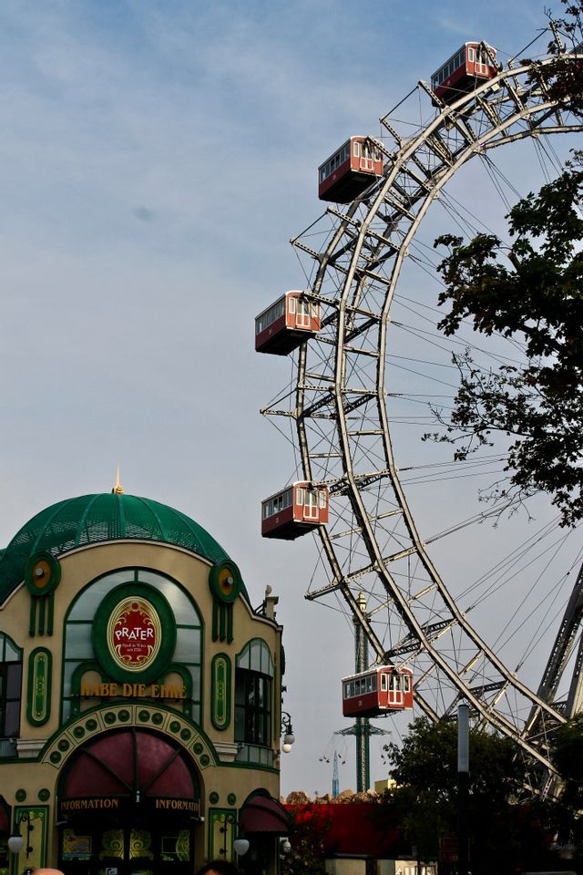Una grande ruota panoramica con cabine rosse si erge accanto a un edificio ornato con una cupola verde sotto un cielo nuvoloso.