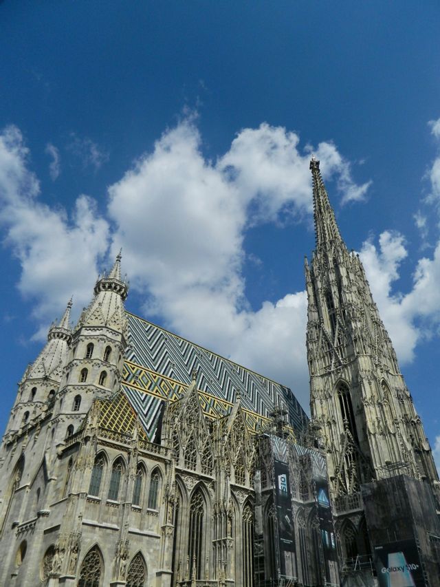Una veduta dal basso di una cattedrale gotica con un tetto in tegole colorate e un'alta guglia contro un cielo azzurro con nuvole.