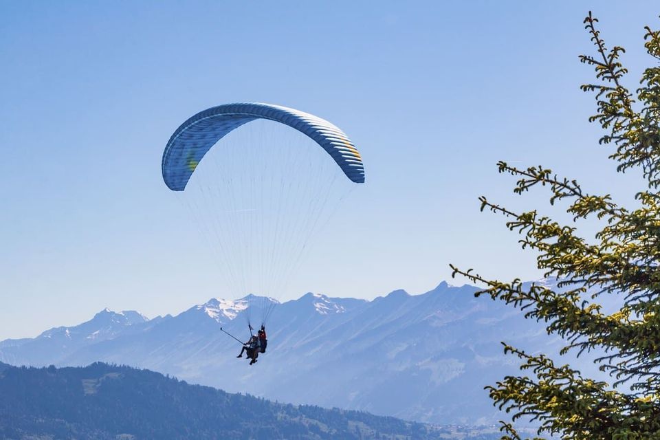 Deux personnes faisant du parapente en tandem avec une perche à selfie au-dessus d'une vaste chaîne de montagnes aux sommets enneigés.