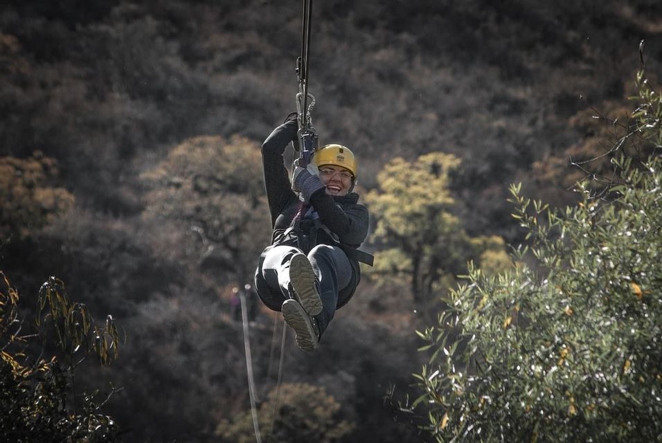 Une femme au casque jaune sourit en glissant sur une tyrolienne, avec un arrière-plan d'arbres flous.
