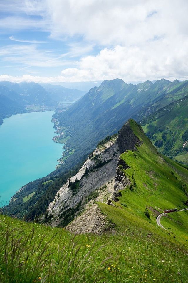 Vue plongeante d'une crête de montagne raide et verte dominant un lac alpin turquoise, avec un train rouge sur une voie sinueuse.